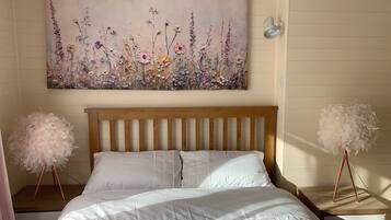 Un dormitorio con cabecera de madera, ropa de cama blanca, una obra de arte floral en la pared y dos mesitas de noche con adornos blancos y suaves.
