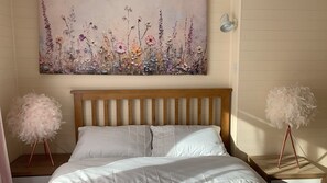 A bedroom with a wooden headboard, white bedding, a floral artwork on the wall, and two bedside tables with fluffy white decorations.