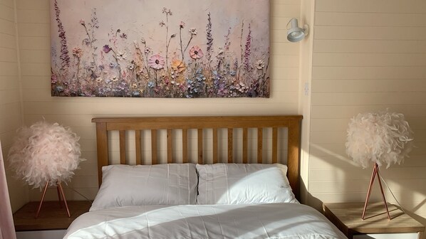 A bedroom with a wooden headboard, white bedding, a floral artwork on the wall, and two bedside tables with fluffy white decorations. - Accessible Mobility Friendly Cottage -garden, Pets (Reydon)