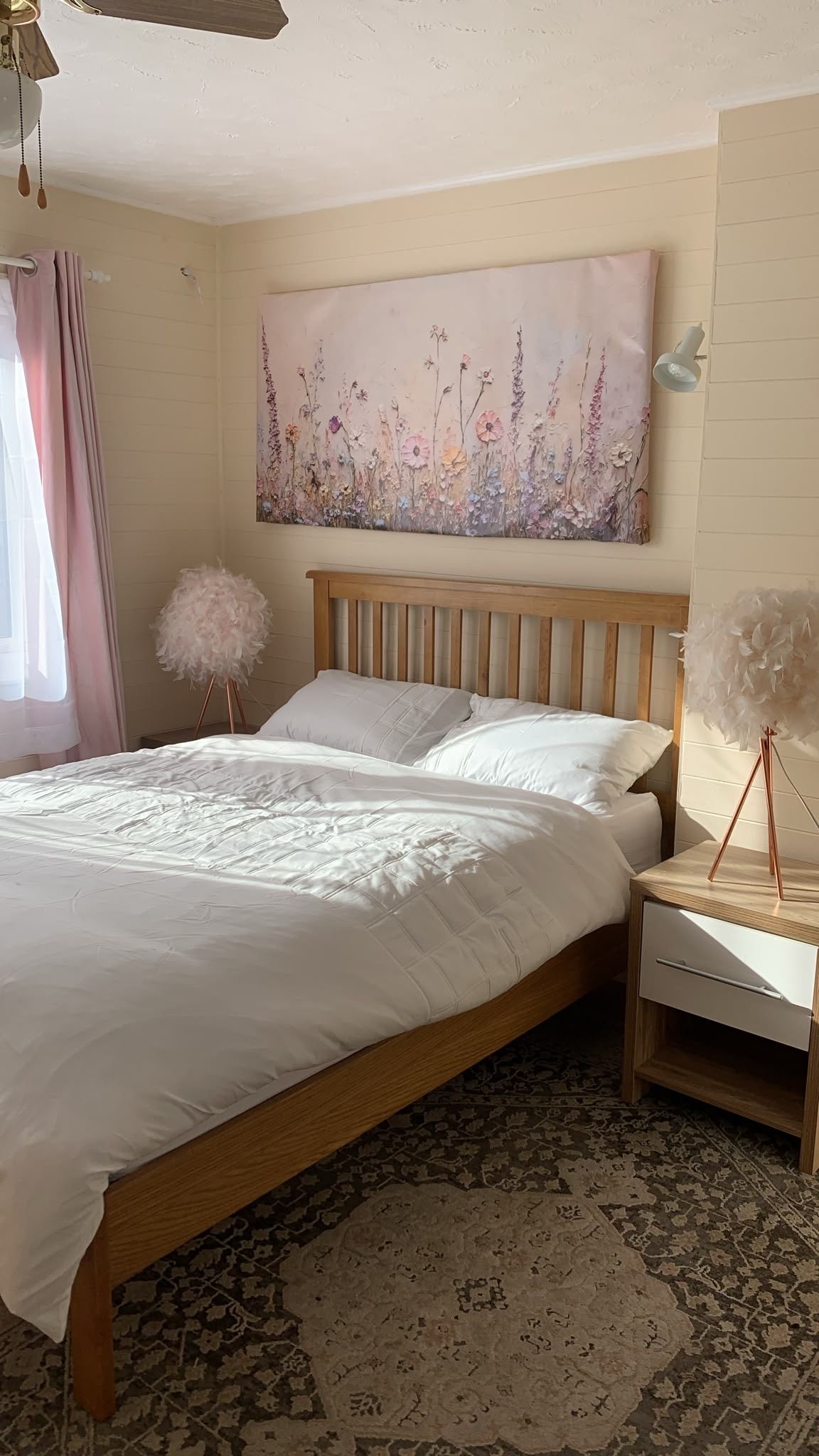 Un dormitorio con cama de madera, ropa de cama blanca, una obra de arte floral en la pared, una mesita de noche con lámpara y una alfombra estampada.