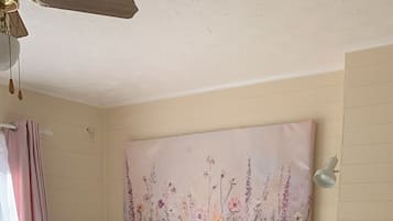 Un dormitorio con cama de madera, ropa de cama blanca, una obra de arte floral en la pared, una mesita de noche con lámpara y una alfombra estampada.
