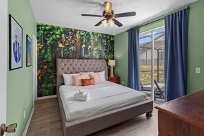 Ein Schlafzimmer mit einem großen Bett, einem Deckenventilator, einem Nachttisch, einer Lampe und Blick ins Freie durch eine Glastür.