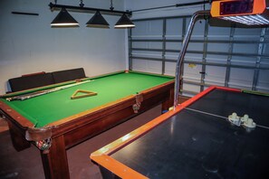 Una mesa de billar con un montón triangular de bolas, tres luces colgantes y una puerta de garaje al fondo.