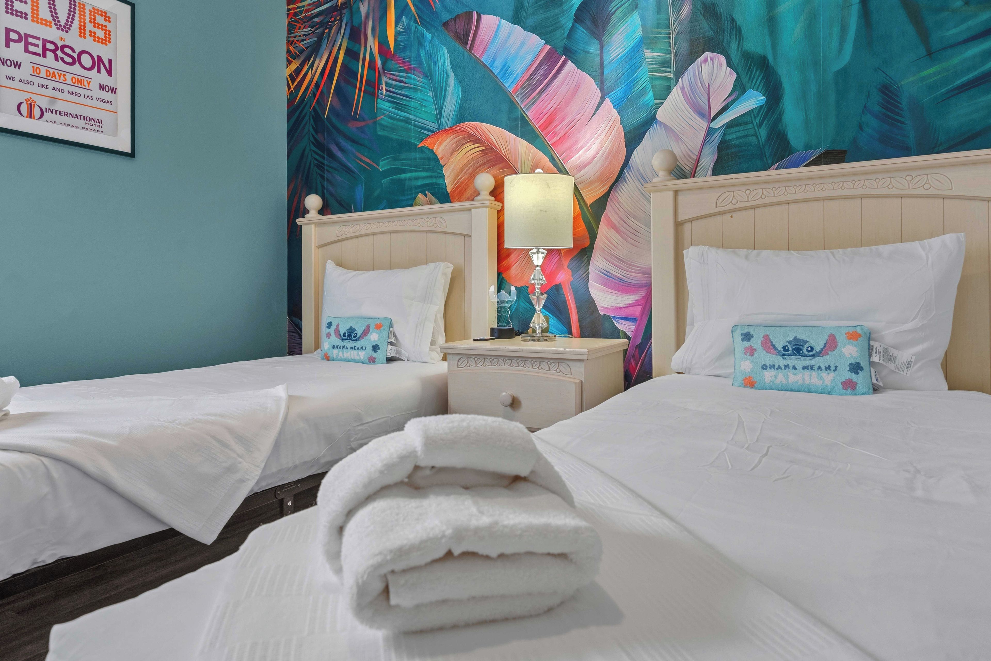 A hotel room with two beds, a nightstand with a lamp, and a tropical-themed wall mural.