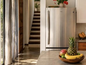 Eine moderne Küche mit einem Edelstahlkühlschrank, einer Holzschale mit Obst auf der Arbeitsfläche und einer Treppe, die zu einer oberen Ebene führt.