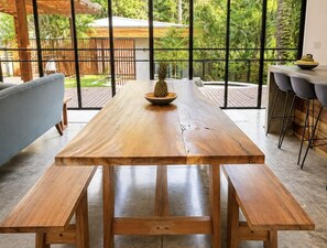 Ein Holztisch mit Bänken, eine Ananas auf dem Tisch und Blick auf einen üppigen Garten durch große Fenster.