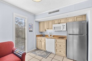 A compact kitchen with a stainless steel refrigerator, a microwave, and a dishwasher. There is a window with blinds, a red chair, and a decorative sign on the wall. - The Pelican Nest-Perfect North Myrtle Beach Getaway (MYRTLE BEACH)