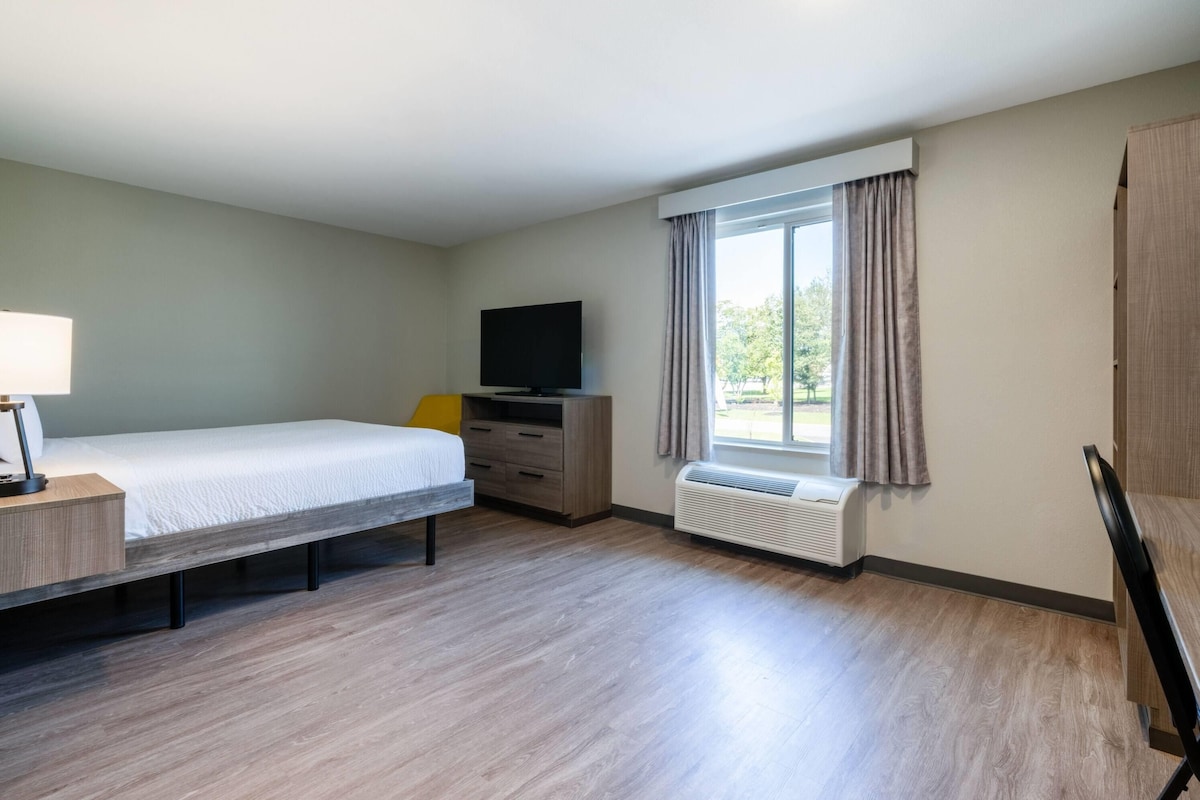 Habitación de hotel con cama, televisión, escritorio con lámpara, calefactor y ventana con cortinas.