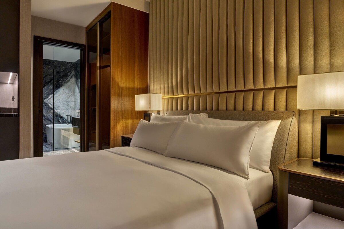A hotel room with a large bed, a wooden headboard, a nightstand with a lamp, and a view of a marble wall and a balcony.