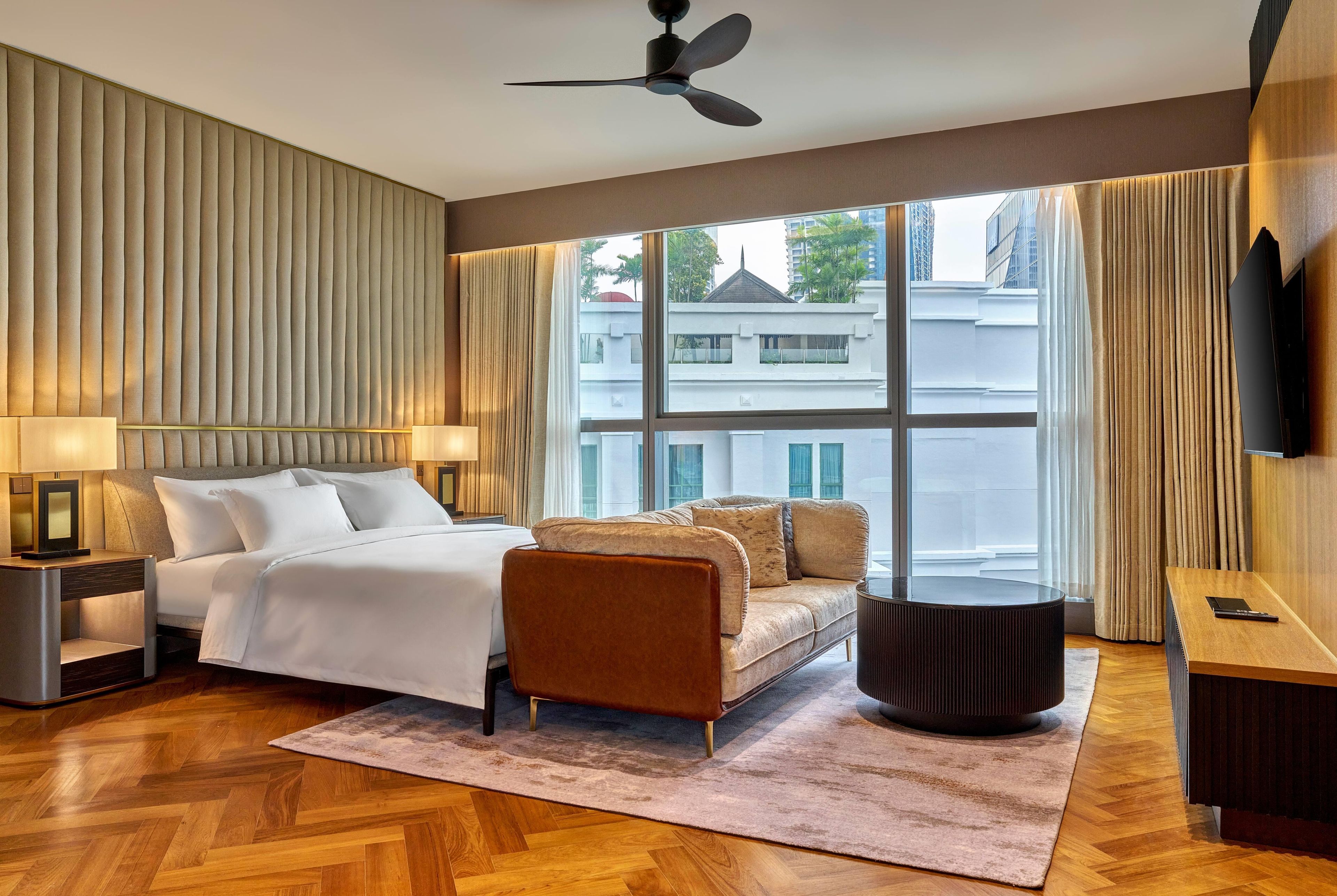 A hotel room with a bed, a sofa, a desk, a television, and a large window with a view of buildings.