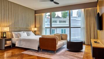A hotel room with a bed, a sofa, a desk, a television, and a large window with a view of buildings.