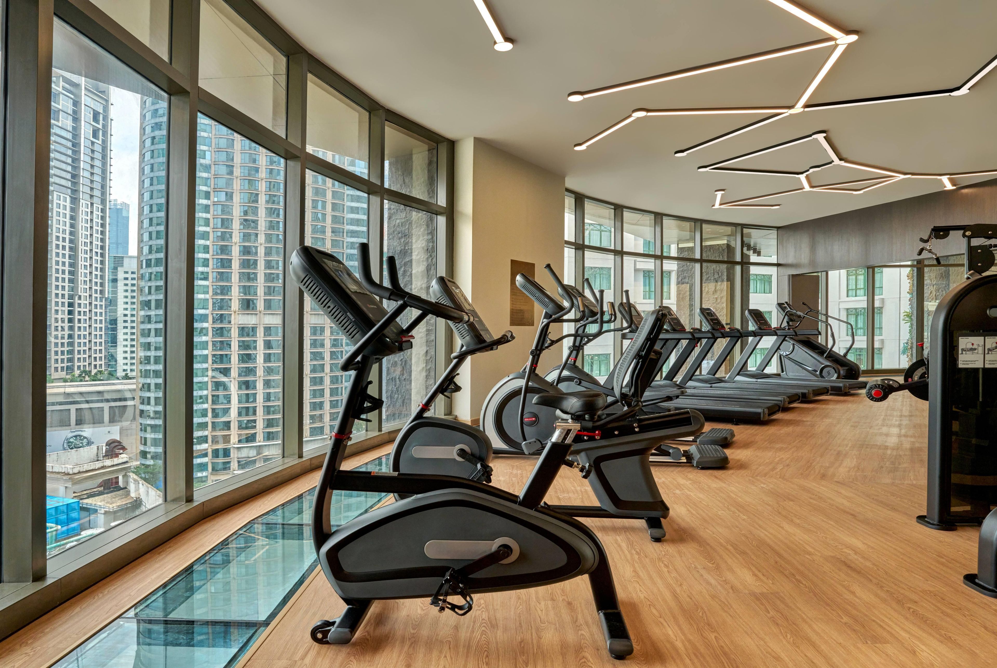 A gym with multiple treadmills and exercise bikes, large windows with a city view, and a modern ceiling design.