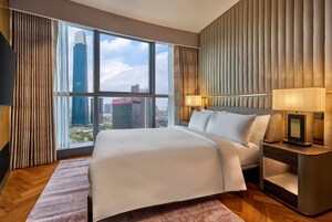 Habitación de hotel con una cama grande, una mesita de noche, una lámpara y vistas a la ciudad a través de amplias ventanas.
