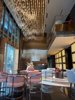 A hotel lobby with a modern chandelier, seating area, reception desk, and large windows.