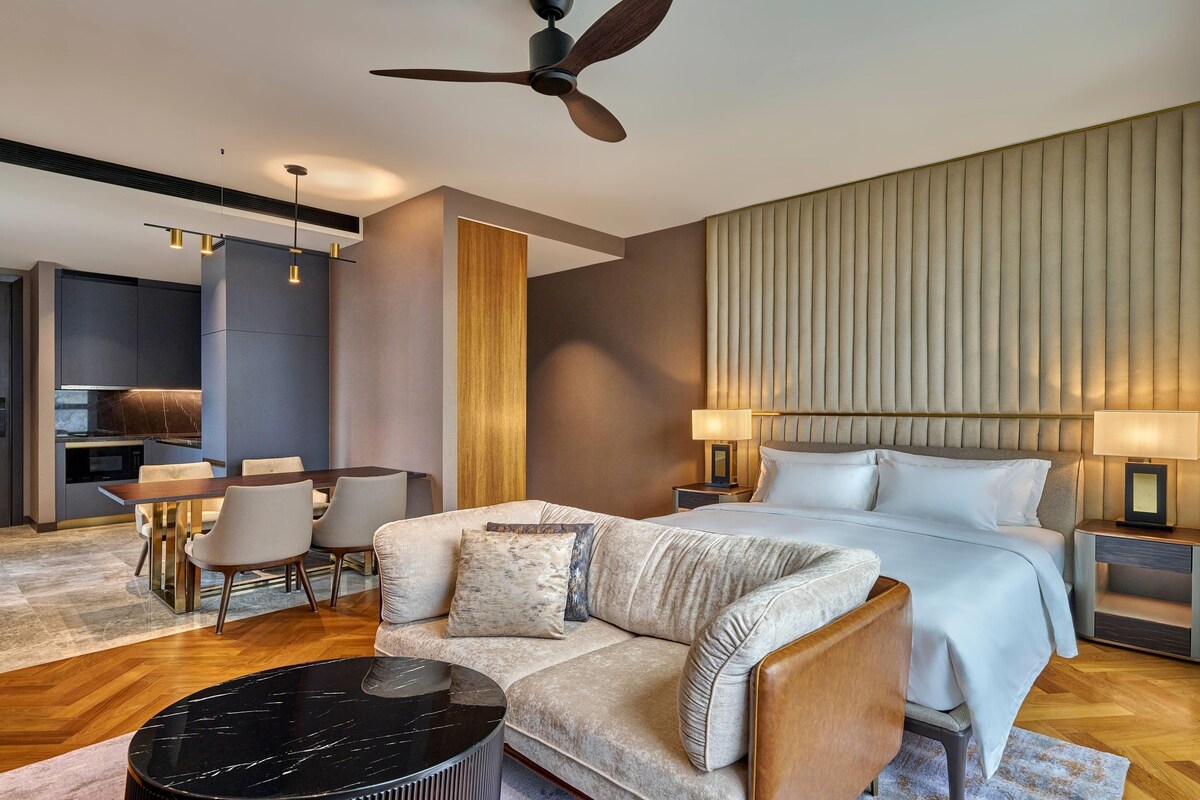 A modern hotel room with a bed, a sofa, a dining table, and a ceiling fan.