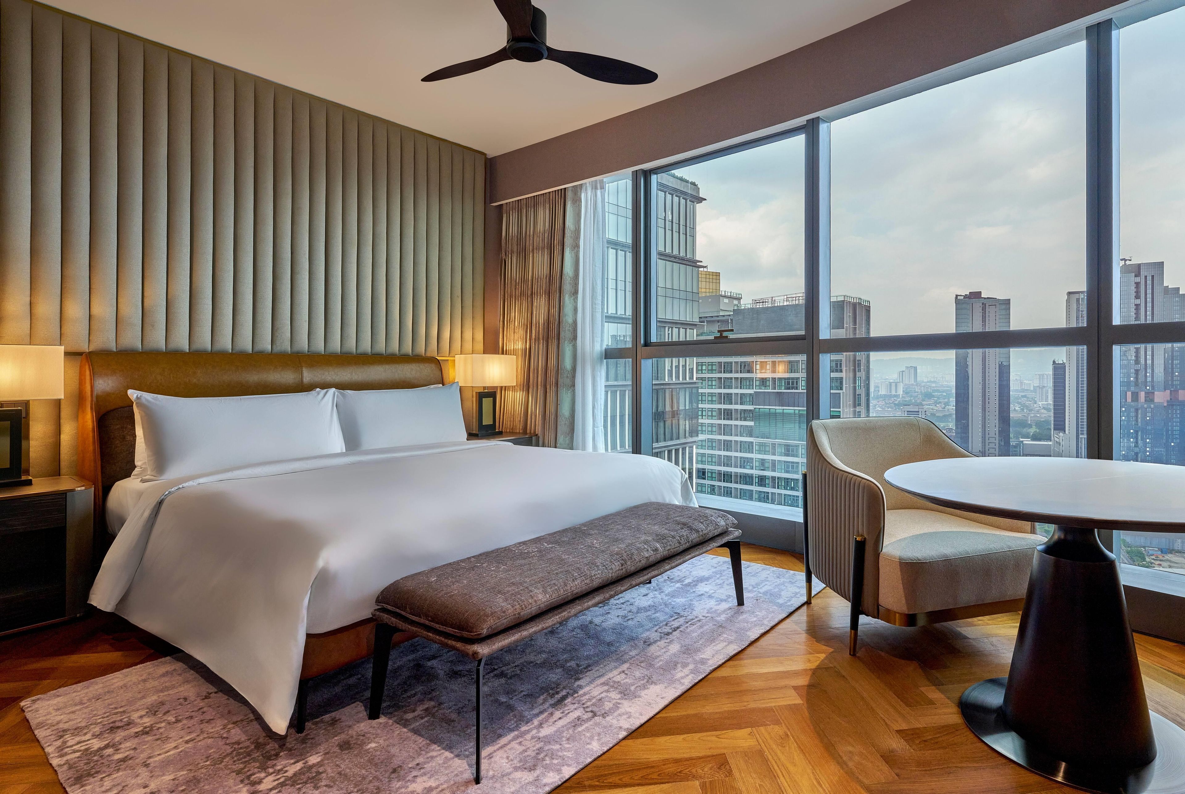 Een hotelkamer met een groot bed, een bankje, een tafel en uitzicht op de stad door grote ramen.