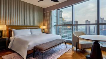 Een hotelkamer met een groot bed, een bankje, een tafel en uitzicht op de stad door grote ramen.