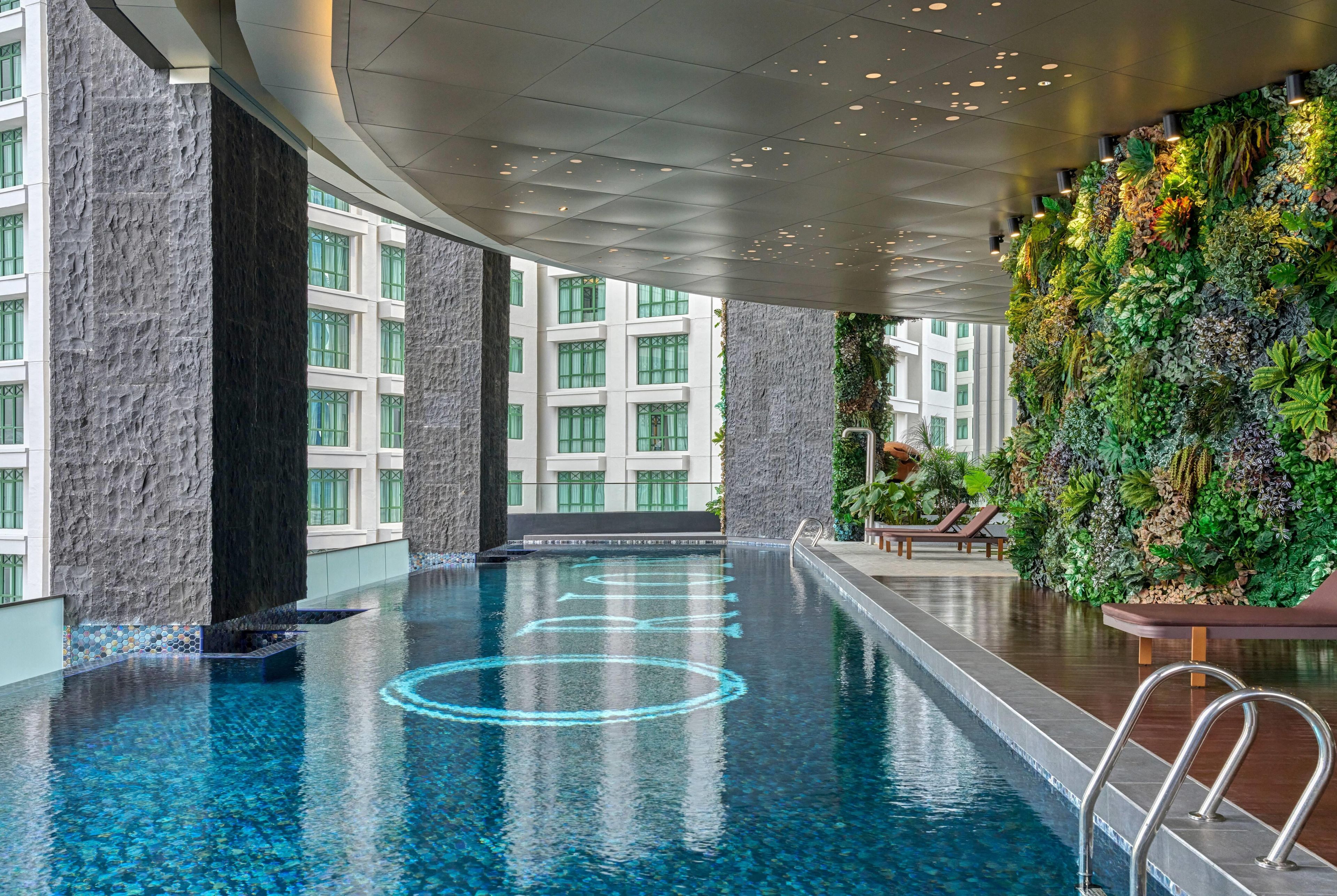 A modern indoor swimming pool with clear blue water, surrounded by stone pillars and a green vertical garden wall.