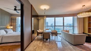 A hotel room with a bed, a dining area with a table and chairs, a sofa, a large window with a city view, and a ceiling fan.