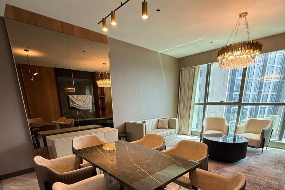 A modern dining area with a rectangular table and six chairs, a chandelier, and a view of the city through large windows.