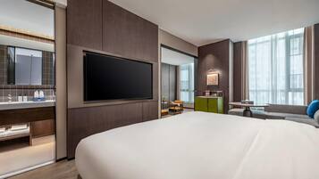 Habitación de hotel moderna con una cama grande, un televisor de pantalla plana montado en la pared, un pequeño comedor y vistas a la ciudad a través de amplias ventanas.