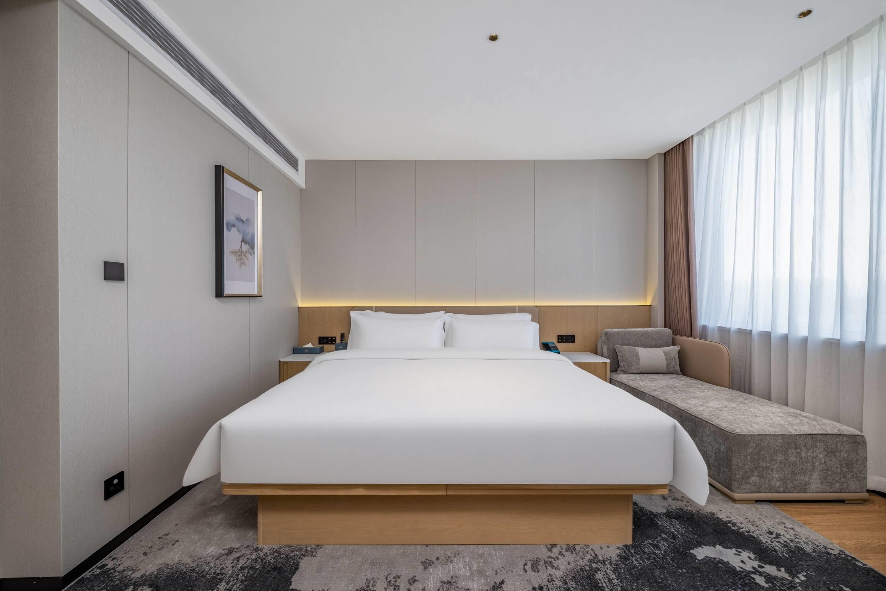 A modern hotel room with a large bed, bedside tables, a sofa, and a large window with curtains.