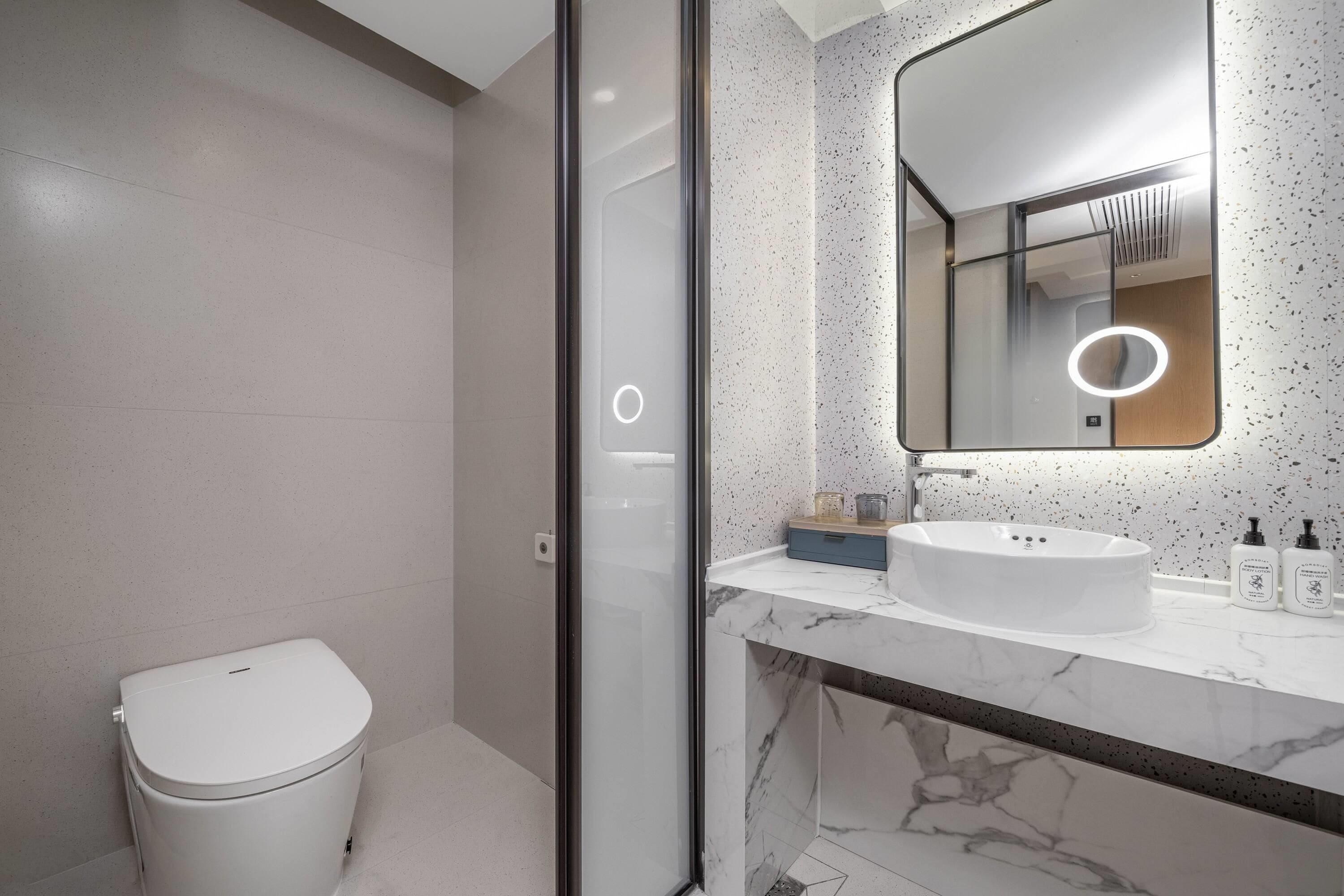 Un baño moderno con inodoro, lavamanos con encimera de mármol y un espejo grande con luz circular.