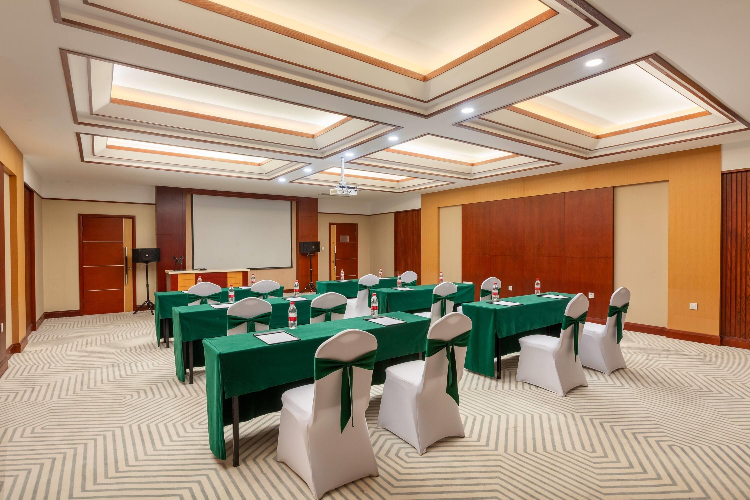 Un salón de conferencias con techo alto, iluminación empotrada y una pantalla para proyector. Mesas dispuestas para una reunión, con sillas y botellas de agua.