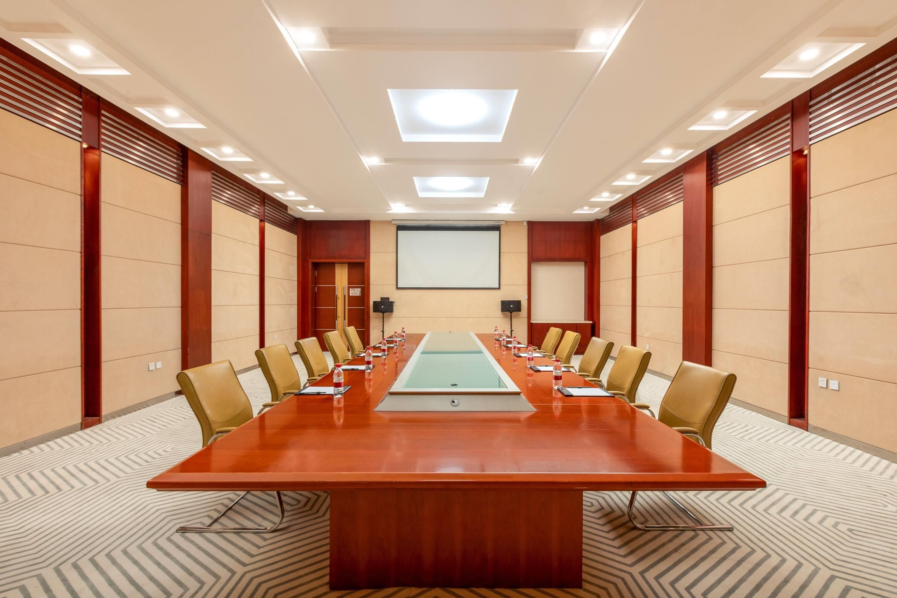 Sala de conferências com uma mesa comprida, cadeiras, tela de projetor e piso coberto de carpete.