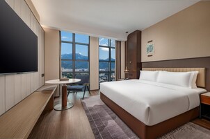 A hotel room with a large bed, a desk with a chair, a TV, and a view of a cityscape through large windows.