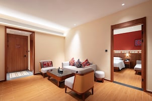 Habitación de hotel con sofá, mesa de centro, silla de mimbre, cama y baño visible a través de una puerta abierta.