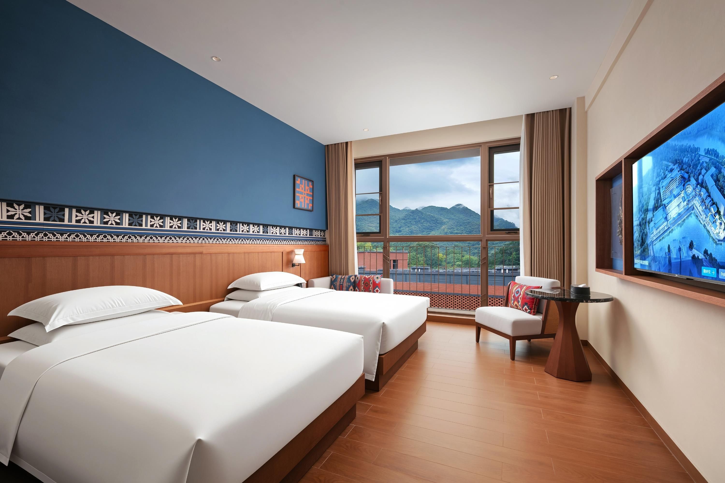 Habitación de hotel con dos camas, un escritorio, una silla y un amplio ventanal con vistas a la montaña.
