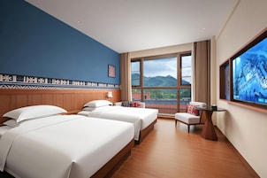 Habitación de hotel con dos camas, un escritorio, una silla y un amplio ventanal con vistas a la montaña.