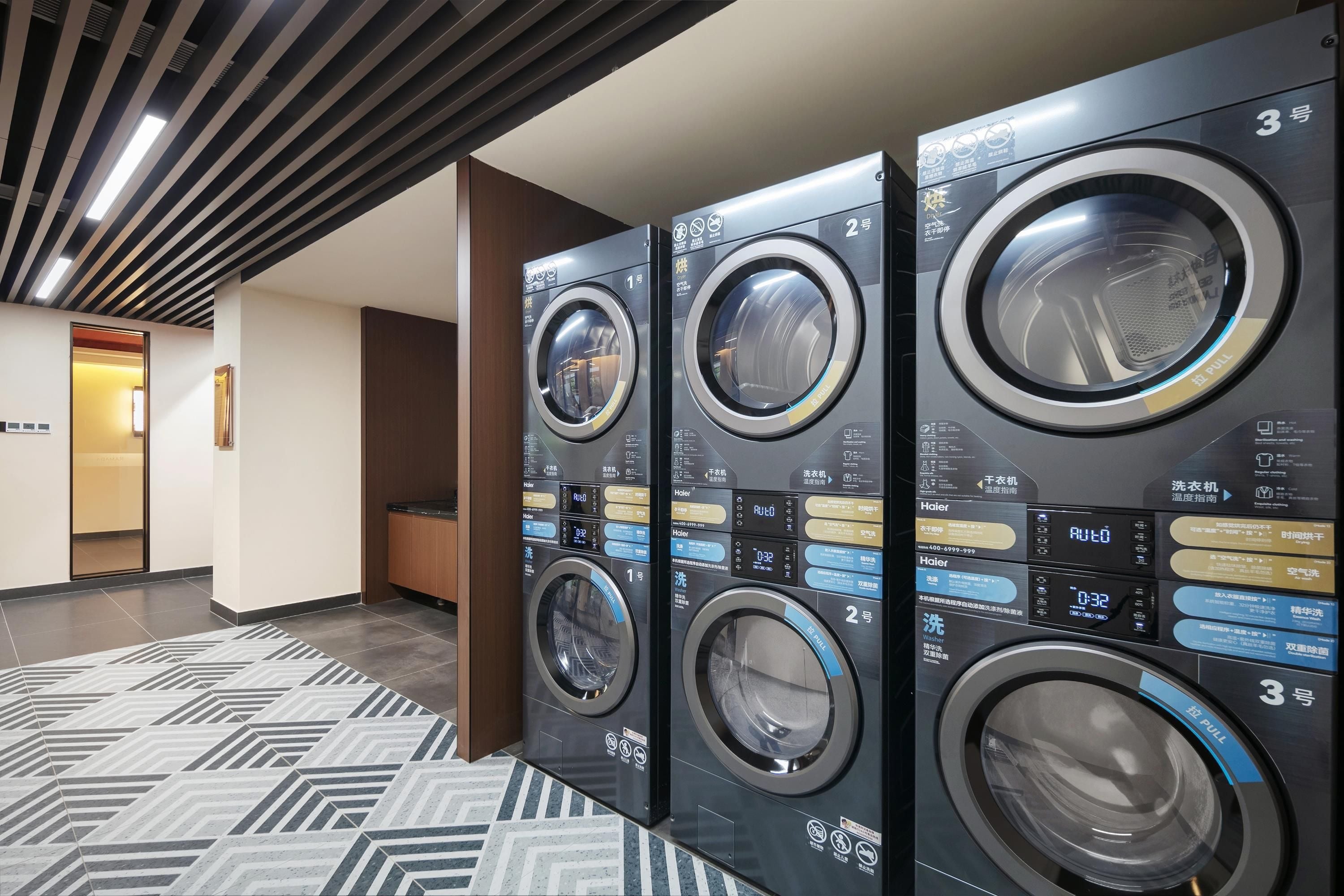 A row of modern, wall-mounted laundry machines with digital displays and control panels, located in a hallway with patterned flooring.