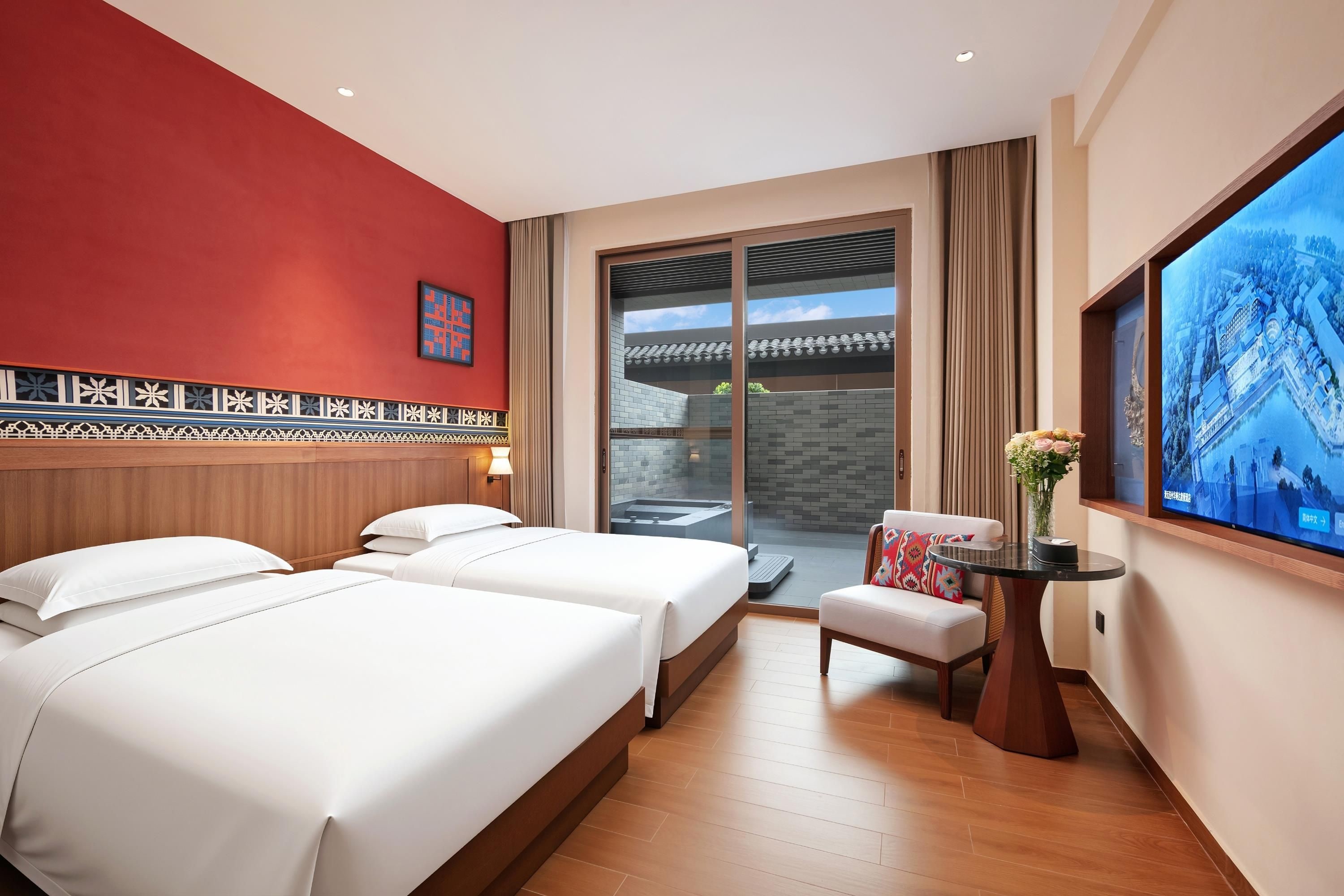 A hotel room with two beds, a small table with a vase, a TV, and a balcony with a view of a building.