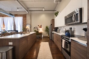 A modern kitchen with a central island, stainless steel appliances, and a brick wall. - The Bentley Collection - The Sinclair (Dallas)