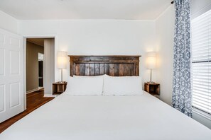 Ein Schlafzimmer mit einem Holzkopfteil, zwei Nachttischlampen, weißer Bettwäsche und einem Fenster mit gemusterten Vorhängen.