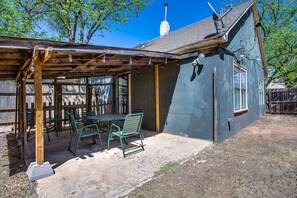 Ein überdachter Patio-Bereich mit Tisch und Stühlen, eine Holz-Pergola und ein Haus mit blauer Fassade.