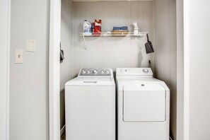 A laundry room with two white washing machines, a shelf with laundry supplies, and a hanging rack. - Brick Road Bungalow 3/1 in South Overton! (Lubbock)