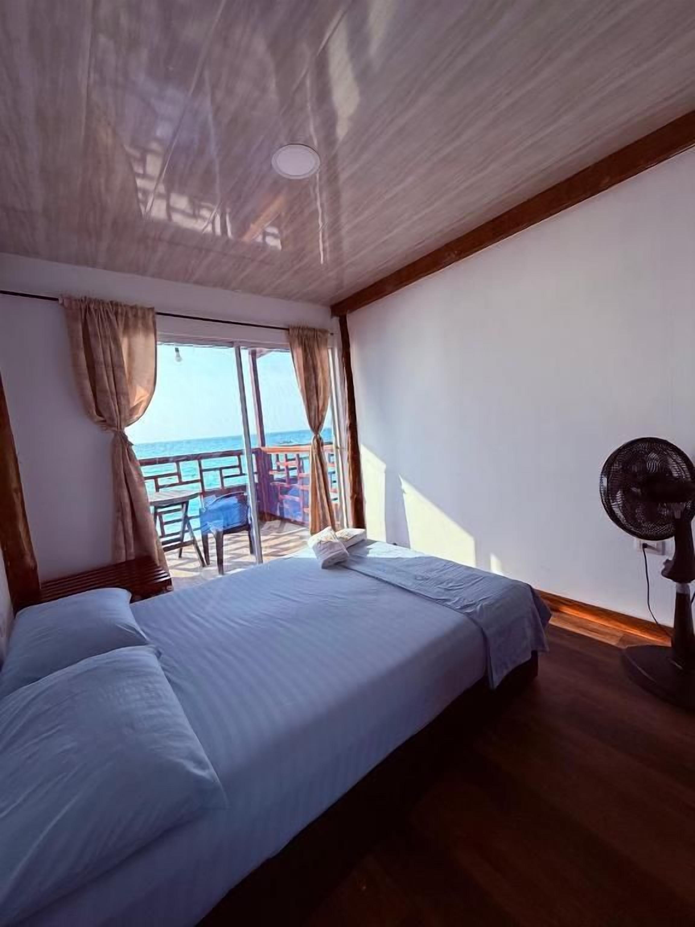 A room with a bed, a fan, and a view of the sea through a window.