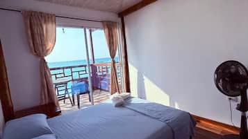 A room with a bed, a fan, and a view of the sea through a window.