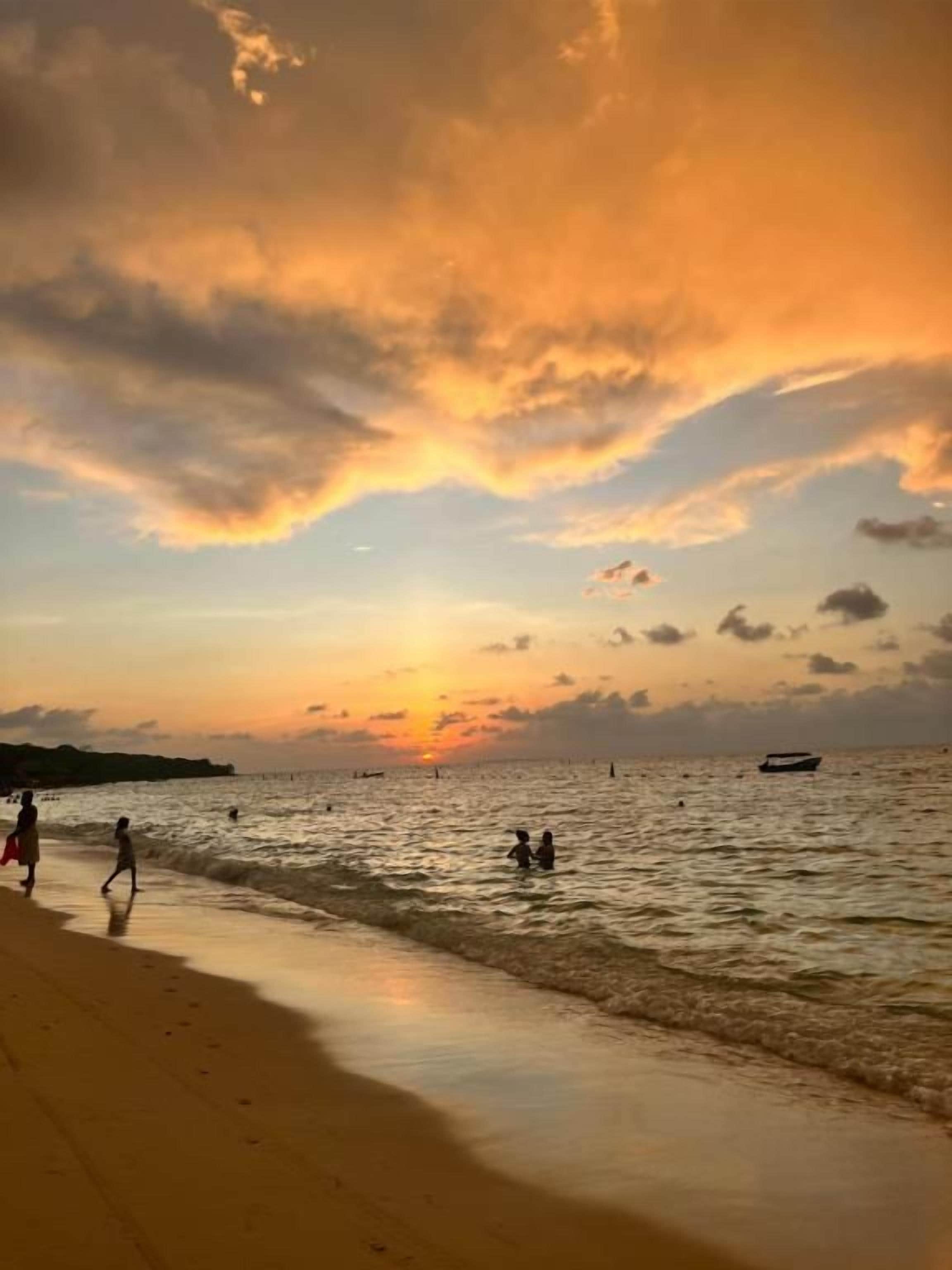 A beach scene with a sunset, people walking along the shore, and a boat in the distance.