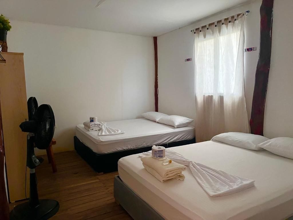 A hotel room with two beds, a wooden floor, a standing fan, and a window with curtains.