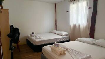 A hotel room with two beds, a wooden floor, a standing fan, and a window with curtains.