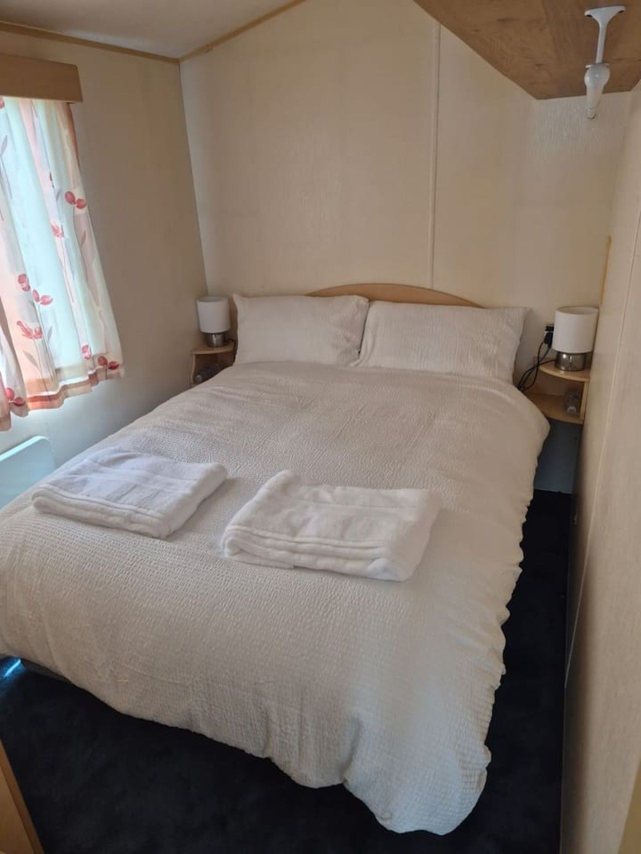 A neatly made bed with a white duvet and two folded towels, a wooden headboard, bedside lamps, and a window with floral curtains.