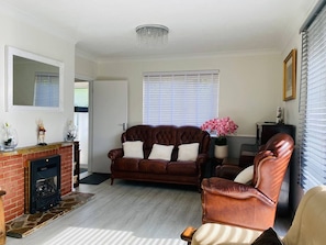 A living room with a fireplace, a brown leather sofa, a leather armchair, and a large window with blinds. - Red Brick Haven 5Bed 2Bath for Memorable Reunions (Isle of Wight)