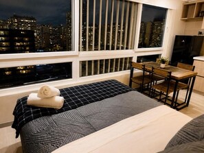Un dormitorio con cama, escritorio y silla. Se ve una ventana con vista a un paisaje urbano de noche.