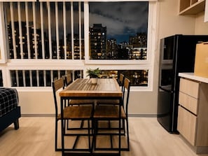 Comedor con vista a un paisaje urbano nocturno, un refrigerador negro y una ventana con persianas.