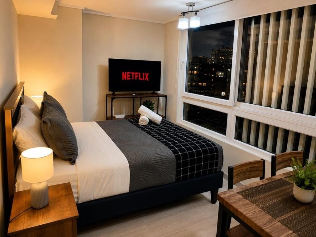 A hotel room with a bed, a TV displaying 'NETFLIX,' a window with blinds, and a table with a plant.