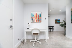 A modern office space with a white desk, a chair, and a painting of people with surfboards. - Central Beach  1-bedroom apartment (Fort Lauderdale)